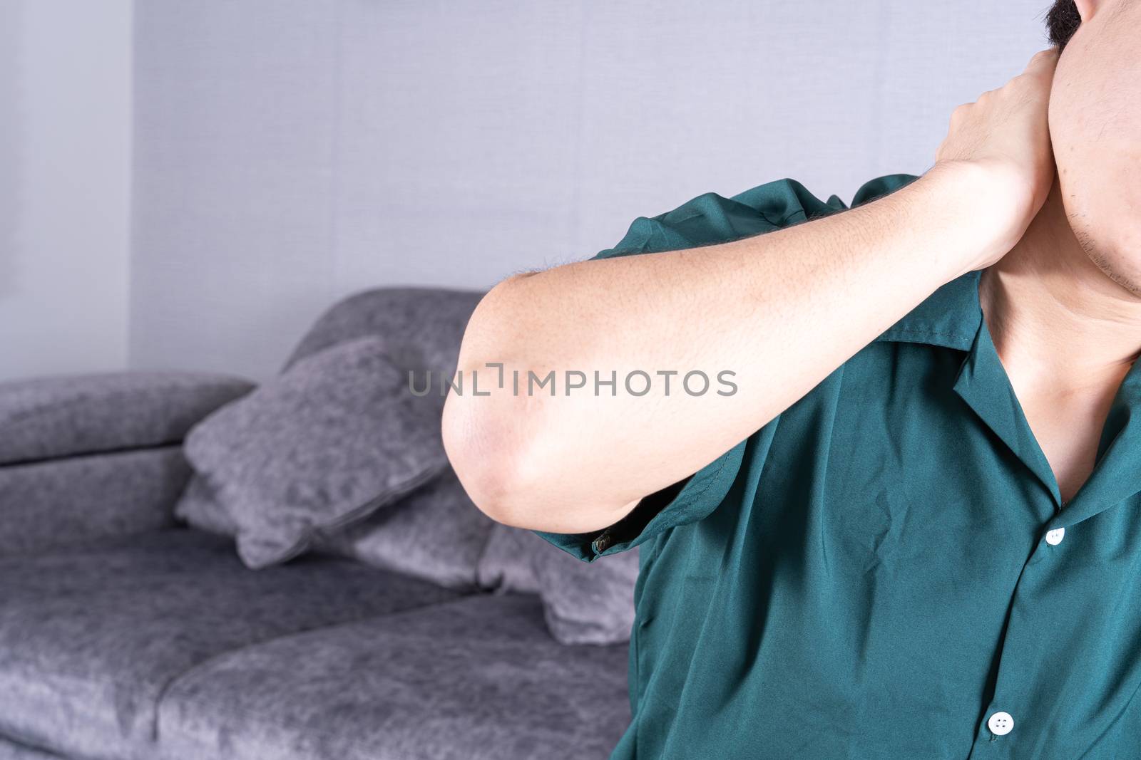 Young man suffering from neck pain while sitting on sofa at home. Healthcare medical or daily life concept.