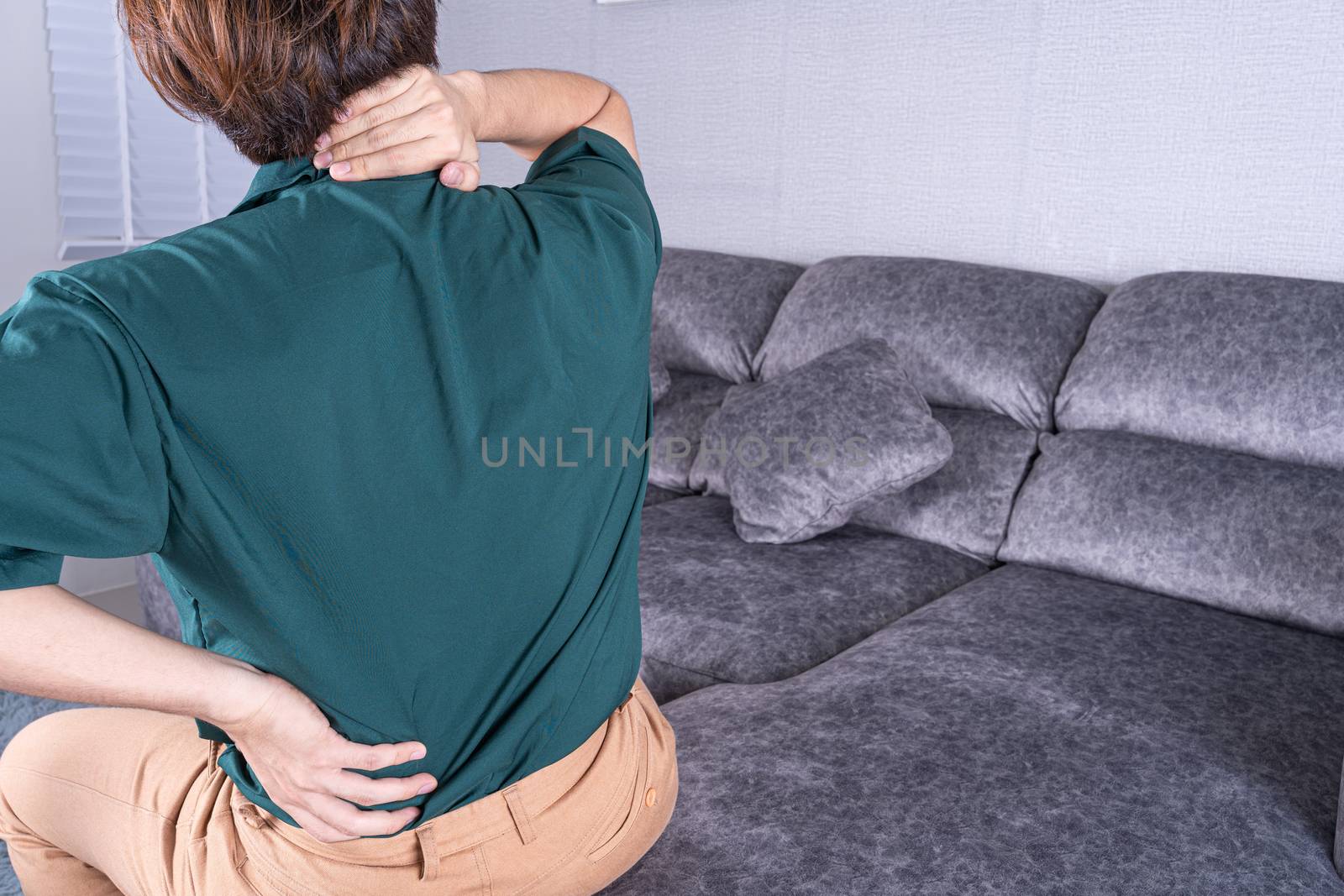 Young man suffering from back and neck pain while sitting on sofa at home. Healthcare medical or daily life concept.