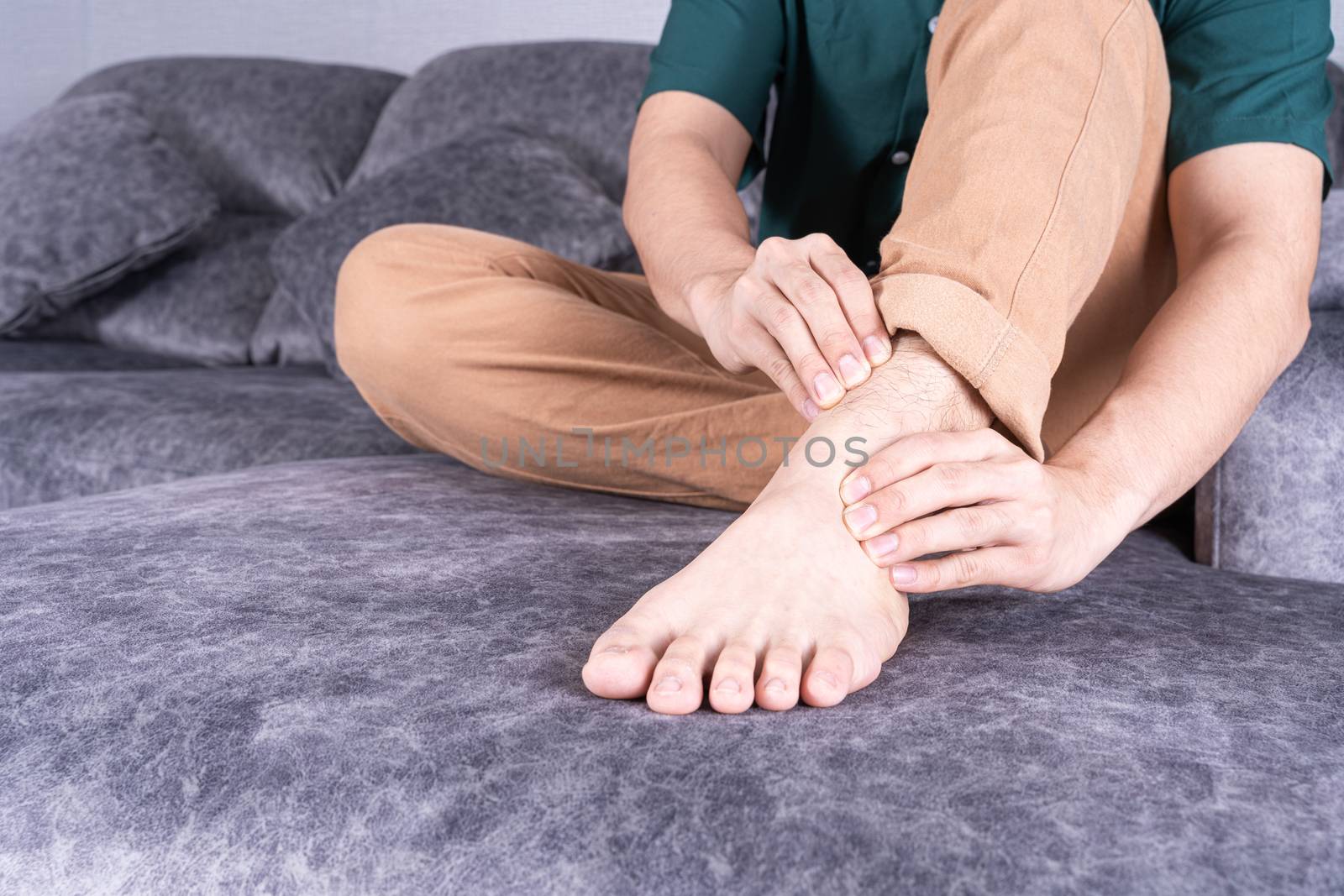 Young man suffering from ankle pain while sitting on sofa at home. Healthcare medical or daily life concept.