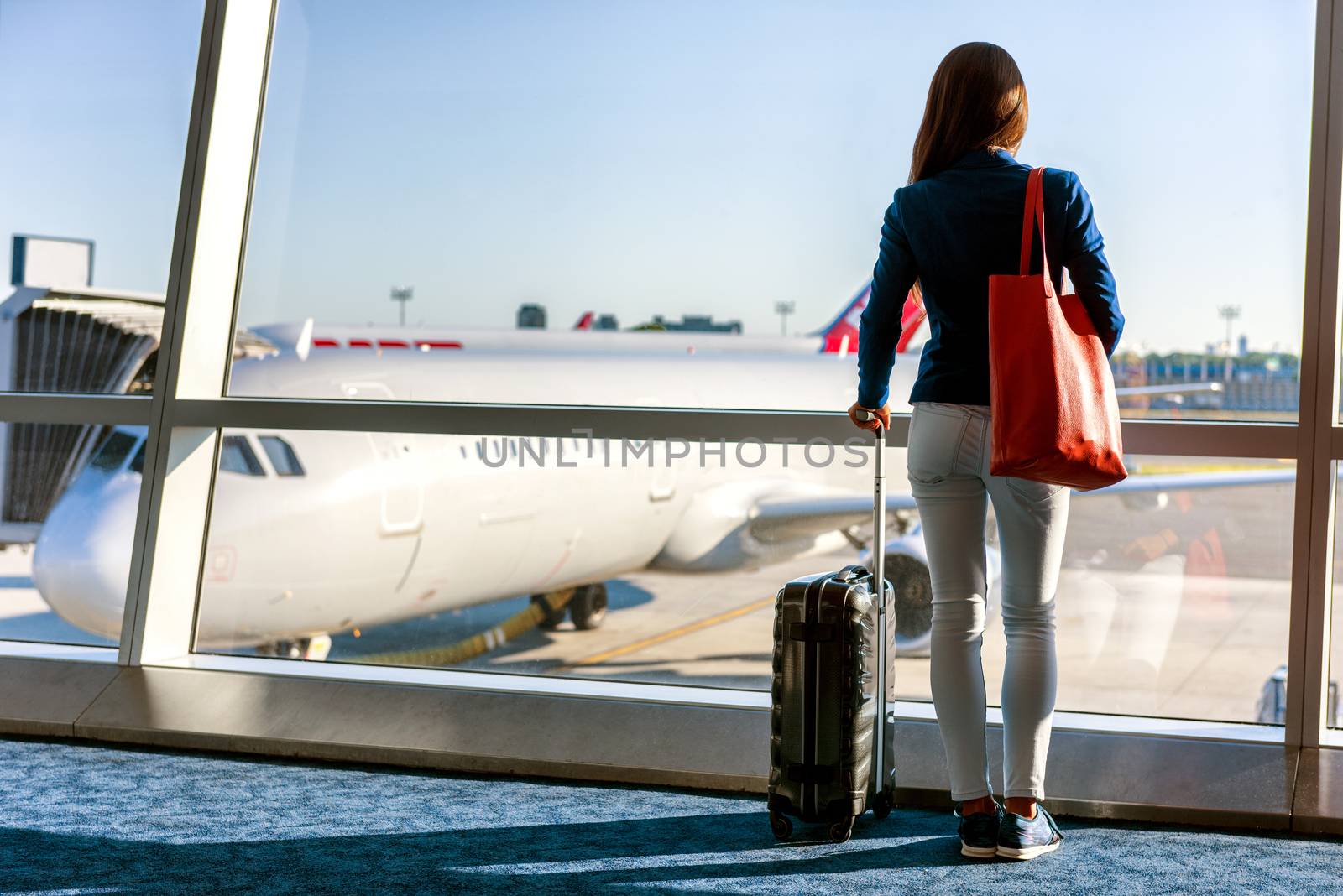 Travel tourist standing with luggages watching sunset at airport window. Unrecognizable woman looking at lounge looking at airplanes while waiting at boarding gate before departure. Travel lifestyle by Maridav