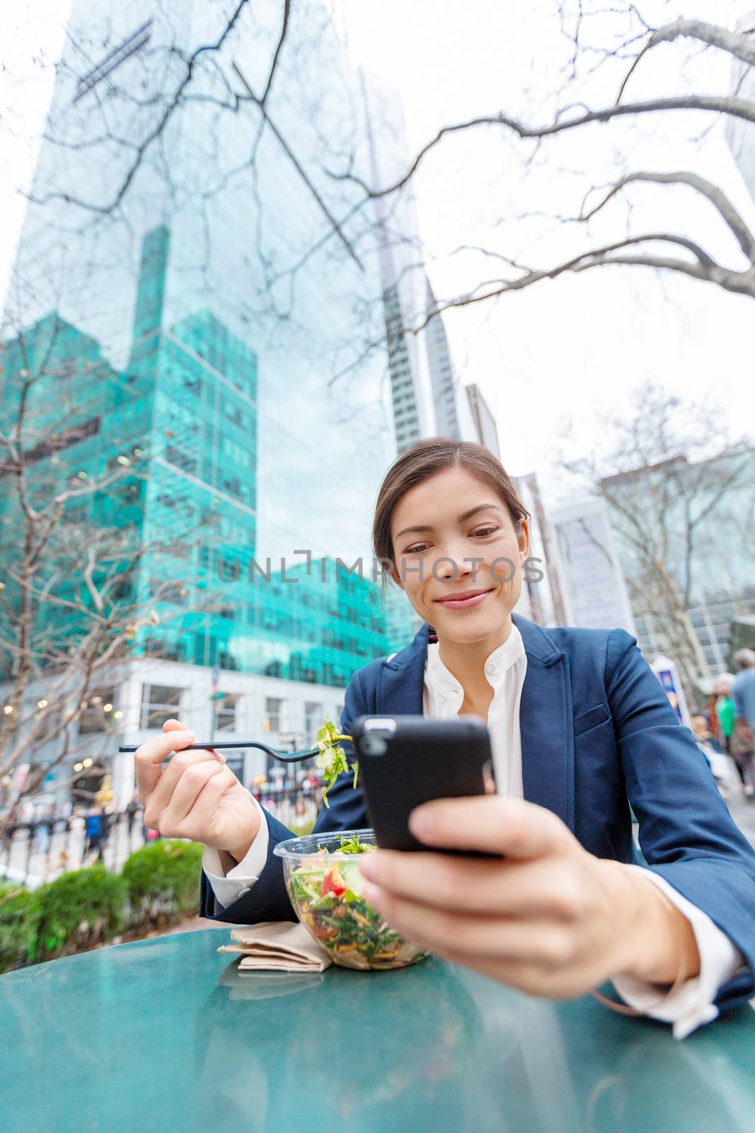 Businesswoman eating salad on lunch break in park. Young professional Asian business woman using phone online data for texting in New York City, USA. Biracial lady Asian Chinese / Caucasian by Maridav