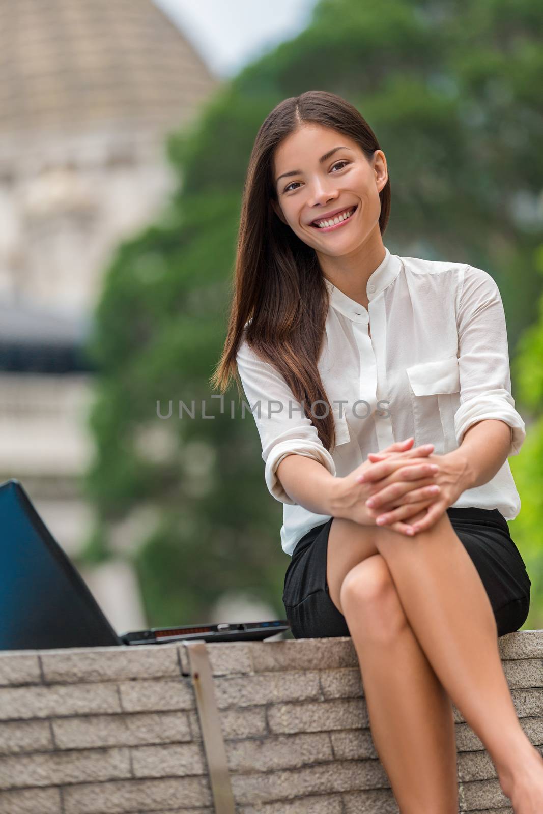 Happy multiracial Asian young woman smiling at camera on work break in city park sitting outdoors. Portrait of Chinese Caucasian girl in her 20s at the office outside, Hong Kong, China, Asia.