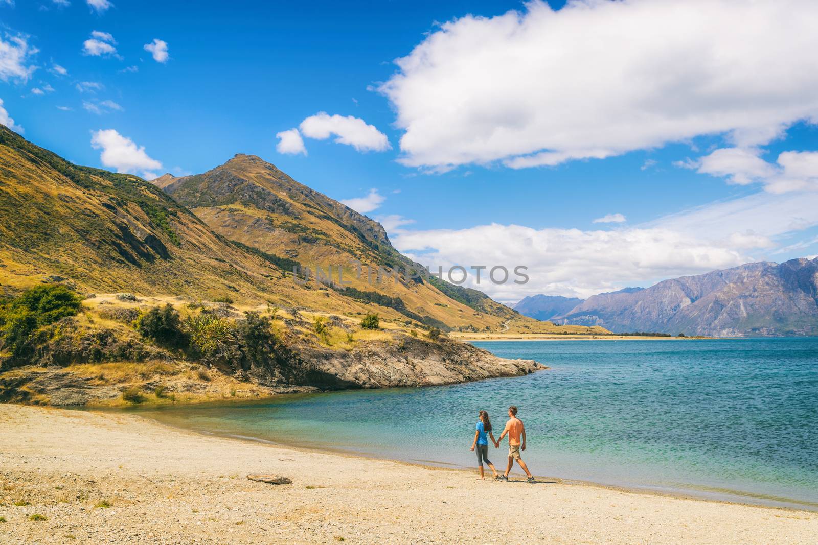 New Zealand travel people tourists visiting Otago region walking on shore of lake Hawea nature landscape. Man and woman happy at beach Near Wanaka. Young tramping hikers adventure lifestyle.