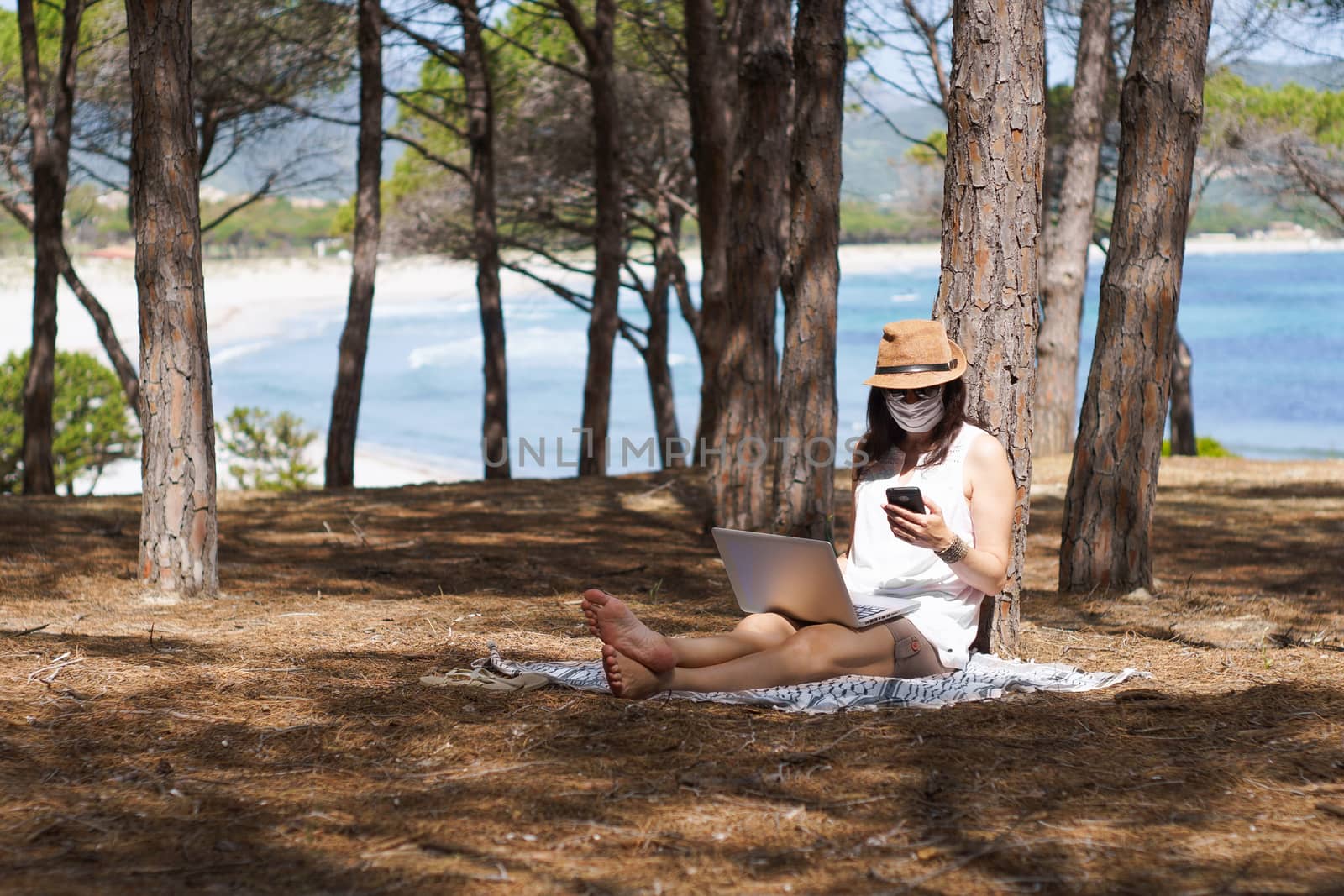 Freelance woman working in vacation on the laptop sitting in a pine forest on the sea with face mask protection for coronavirus, smart working lifestyle concept theme and life choice