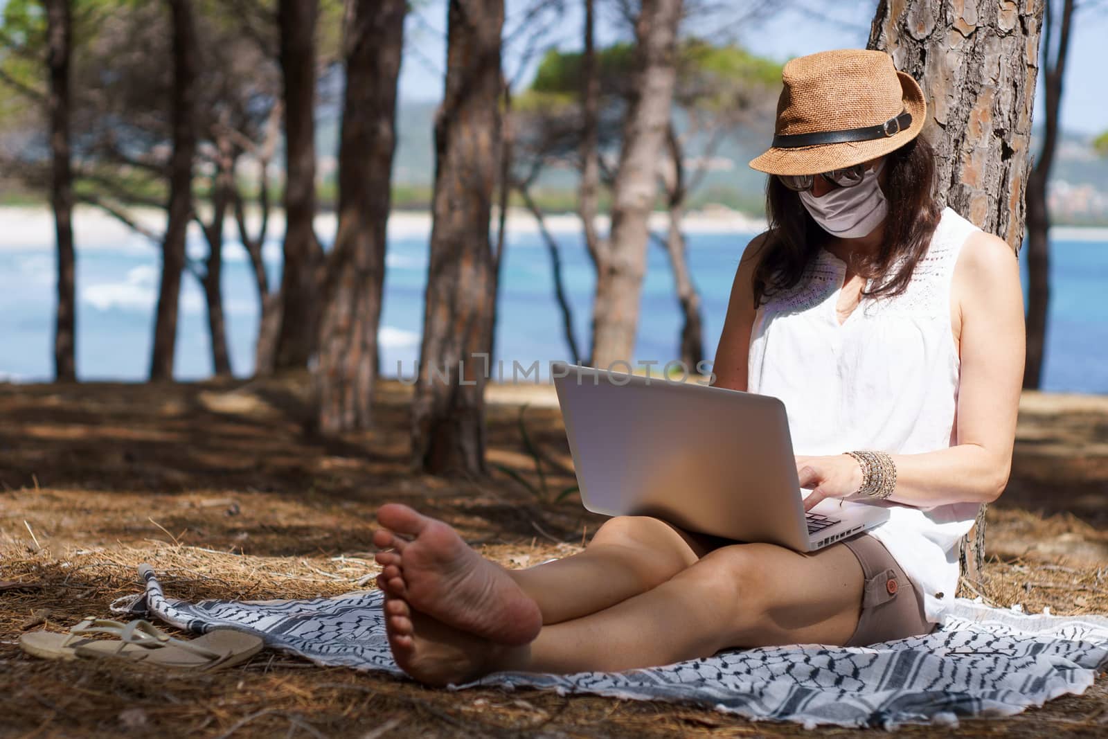 Freelance woman working in vacation on the laptop sitting in a pine forest on the sea with face mask protection for coronavirus, smart working lifestyle concept theme and life choice by robbyfontanesi