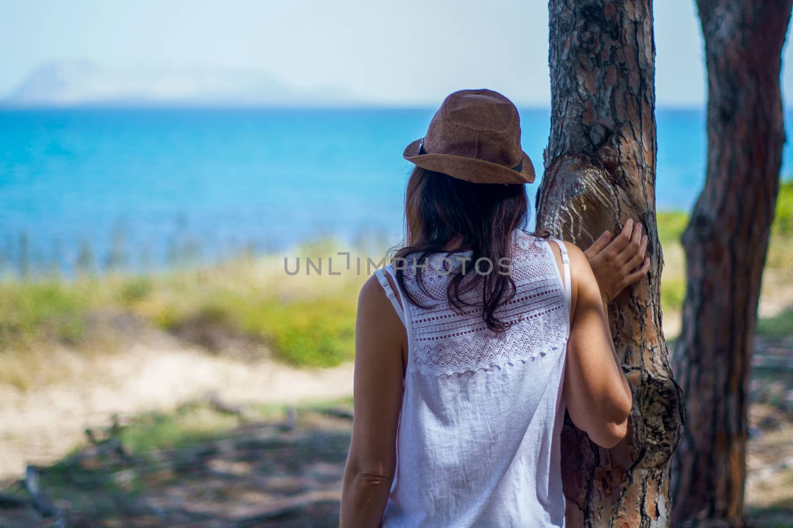 Pensive young woman with long hair and hat, seen from behind, looks at the sea leaning against a tree in a pine forest by robbyfontanesi