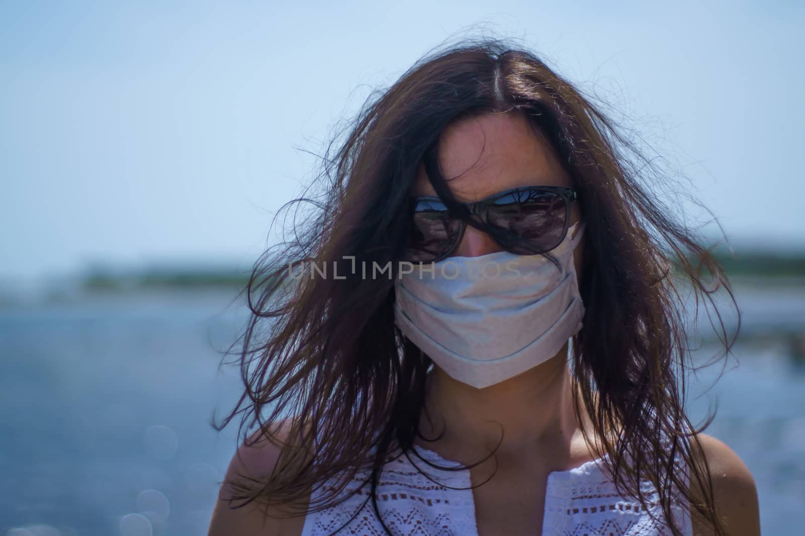 Coronavirus seaside holidays: half-length shot of a woman at the beach look at the camera with the mask for Covid-19 pandemic with cloudy sky