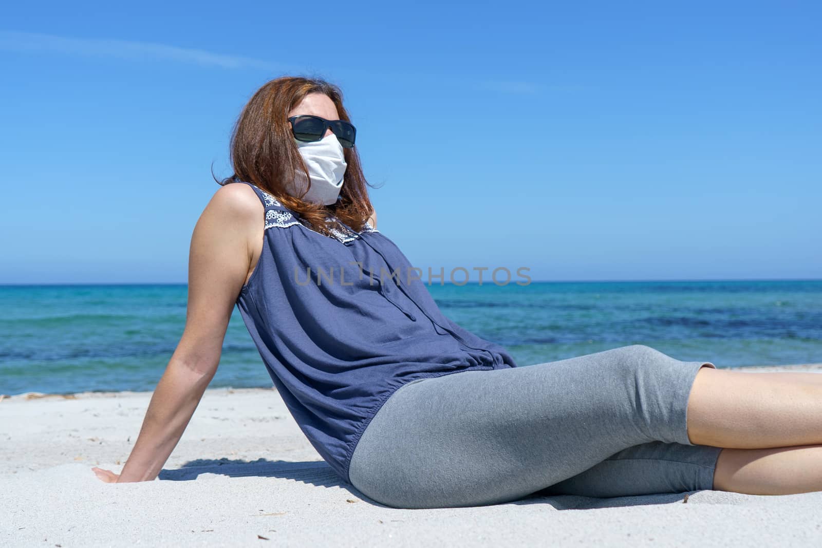 Coronavirus seaside holidays: a woman sitting on the sand at the beach look at the sun with the mask for Covid-19 pandemic