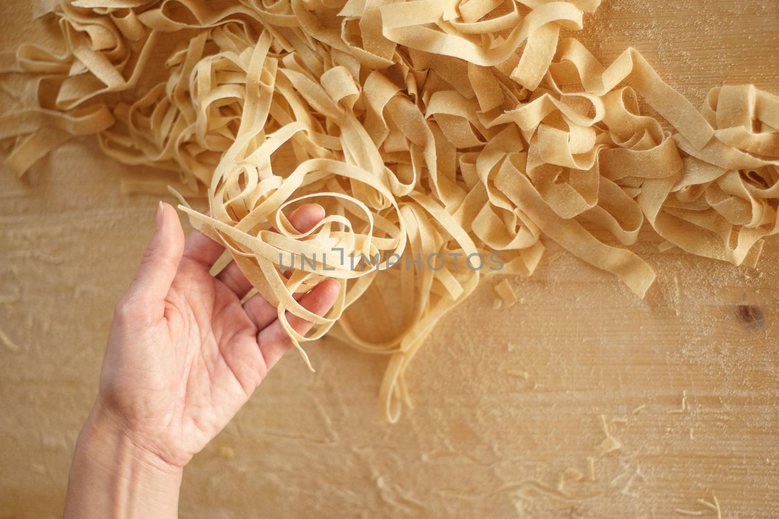 Preparing fresh homemade pasta: close-up view of woman's hand pulling up some fresh tagliatelle on wooden work table in natural sunlight by robbyfontanesi