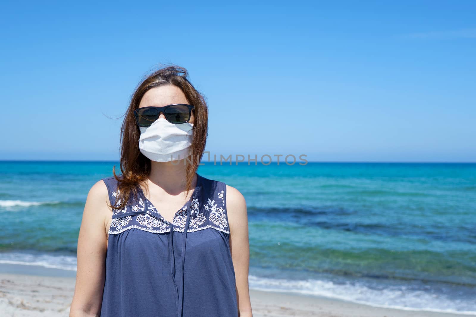 Coronavirus seaside holidays: a woman standing on the sand at the beach looking at the camera with the mask for Covid-19 pandemic by robbyfontanesi