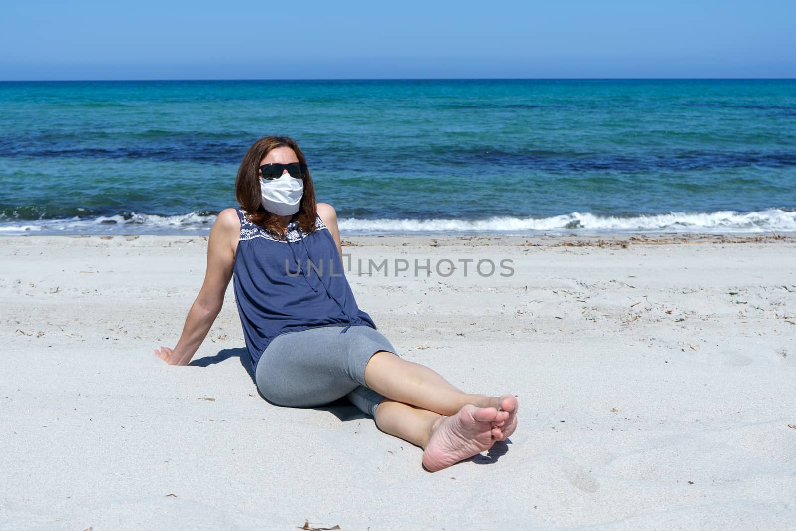 Coronavirus seaside holidays: a woman sitting on the sand at the beach look at the sun with the mask for Covid-19 pandemic by robbyfontanesi