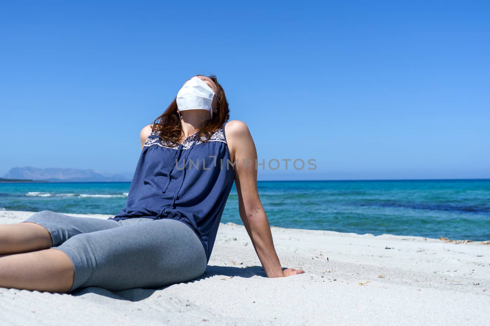 Coronavirus seaside holidays: a woman sitting on the sand at the beach look at the sun with the mask for Covid-19 pandemic by robbyfontanesi