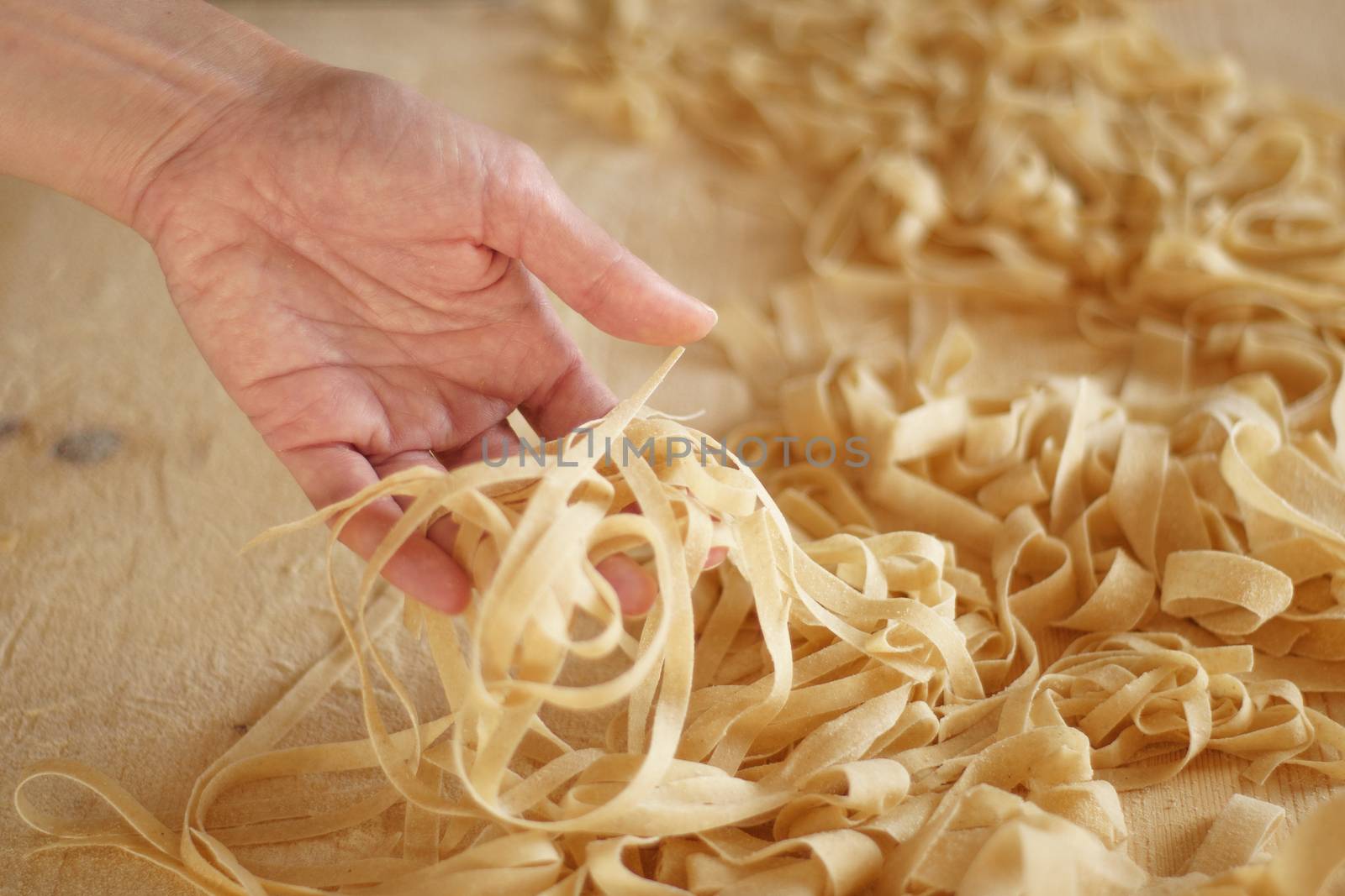 Preparing fresh homemade pasta: close-up view of woman's hand pulling up some fresh tagliatelle on wooden work table in natural sunlight