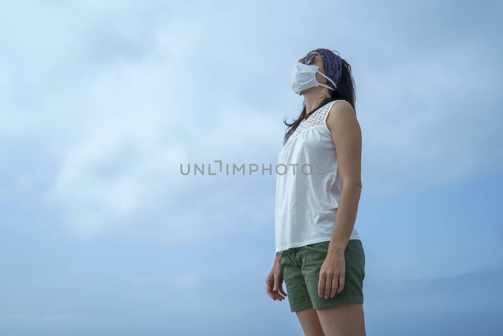 Coronavirus seaside holidays: shot of a woman at the beach looking at the sun with the mask for Covid-19 pandemic with cloudy sky by robbyfontanesi