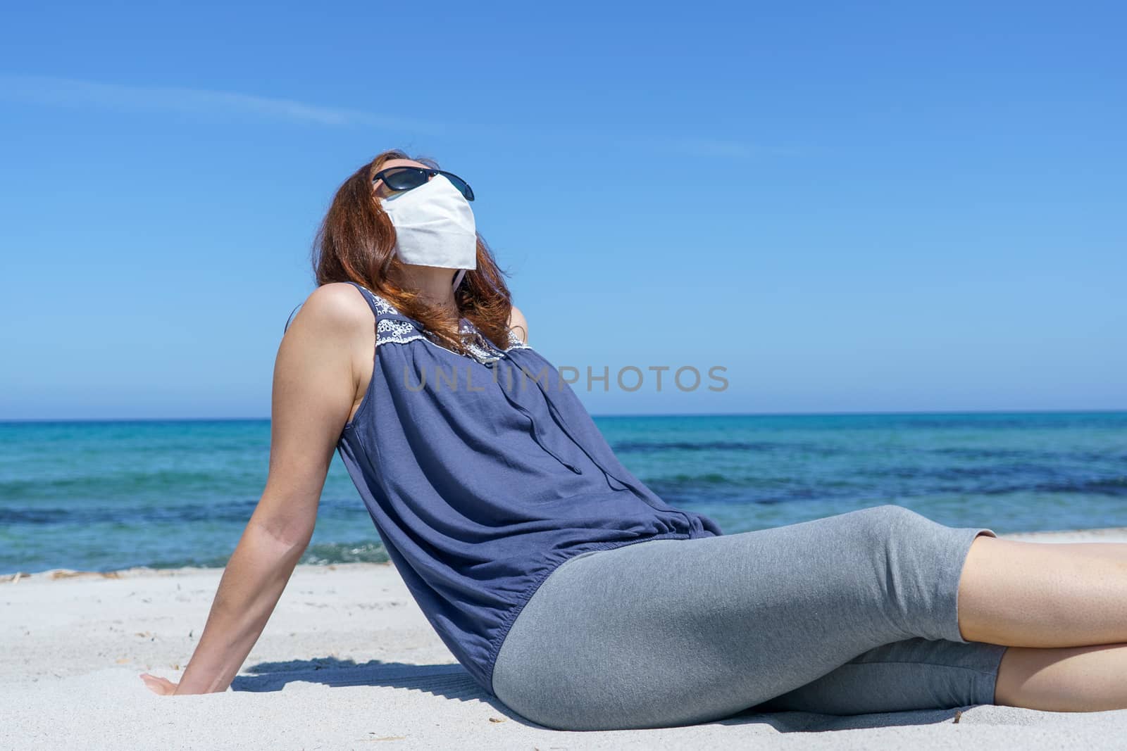 Coronavirus seaside holidays: a woman sitting on the sand at the beach look at the sun with the mask for Covid-19 pandemic by robbyfontanesi