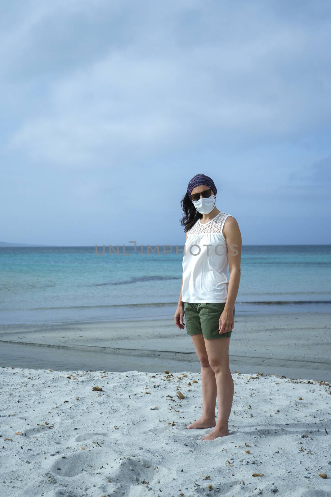 Coronavirus seaside holidays: shot of a woman at the beach look at the camera with the mask for Covid-19 pandemic with cloudy sky by robbyfontanesi