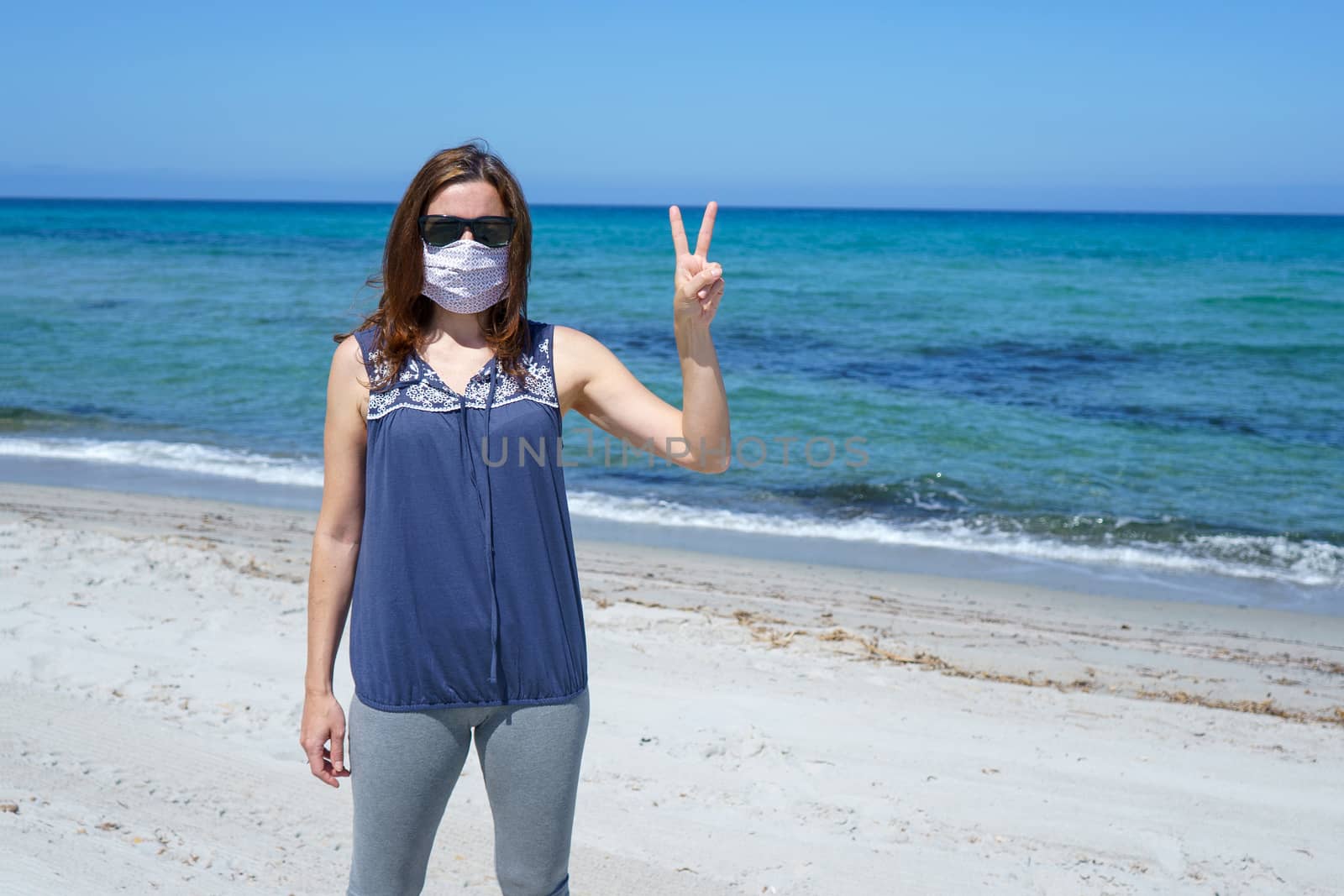 Coronavirus seaside holidays: a woman standing on the sand at the beach looking at the camera with fingers in victory and the mask for Covid-19 pandemic