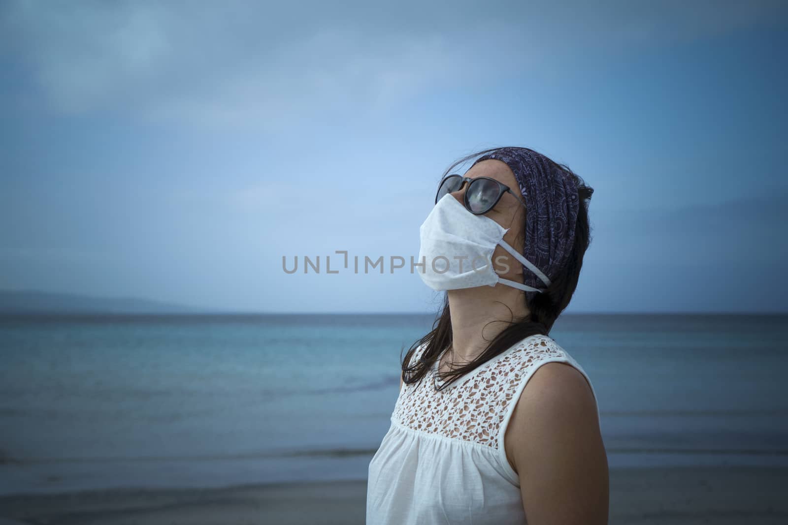 Coronavirus seaside holidays: half-length shot of a woman at the beach looking at the sun with the mask for Covid-19 pandemic by robbyfontanesi
