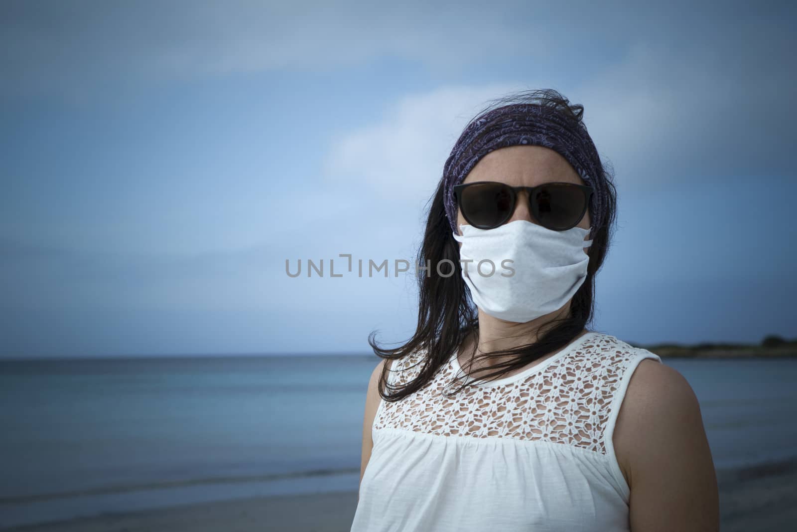 Coronavirus seaside holidays: half-length shot of a woman at the beach look at the camera with the mask for Covid-19 pandemic with cloudy sky by robbyfontanesi