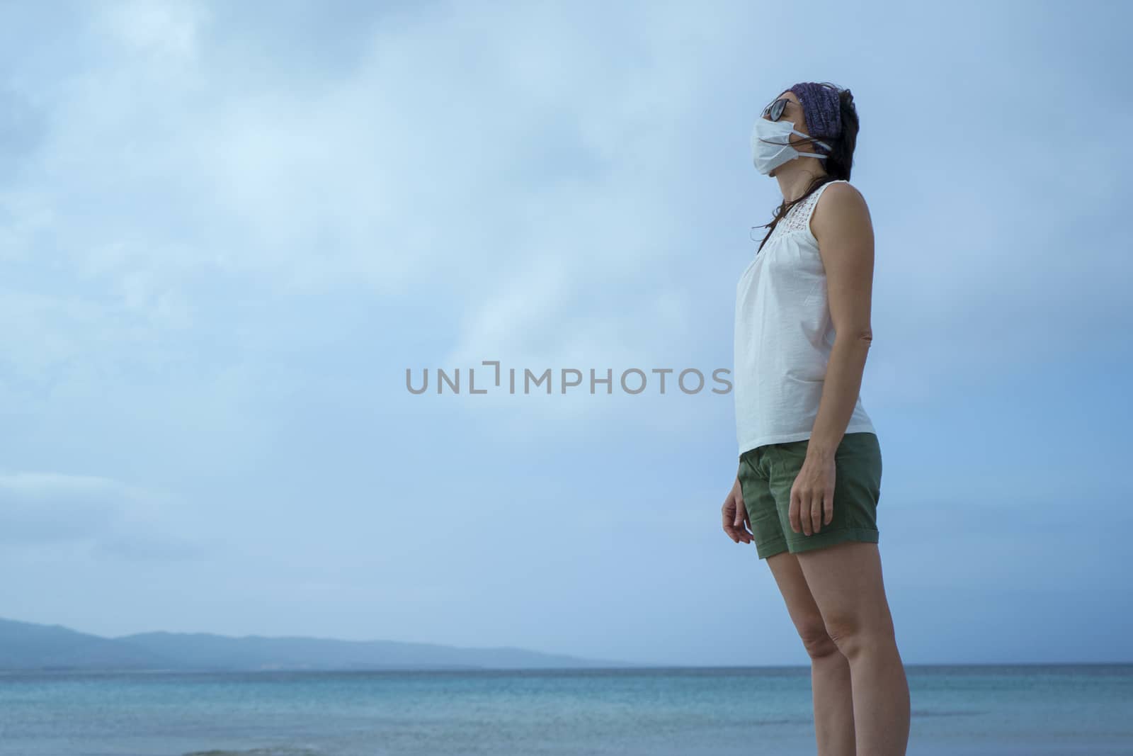 Coronavirus seaside holidays: shot of a woman at the beach looking at the sun with the mask for Covid-19 pandemic with cloudy sky