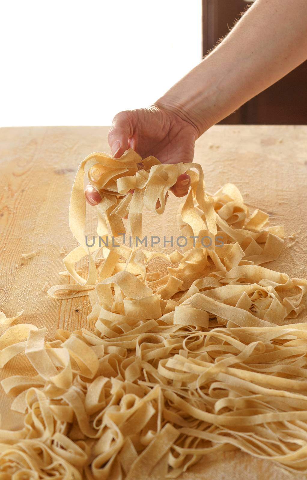 Preparing fresh homemade pasta: close-up view of woman's hand pulling up some fresh fettuccine on wooden work table in natural sunlight by robbyfontanesi