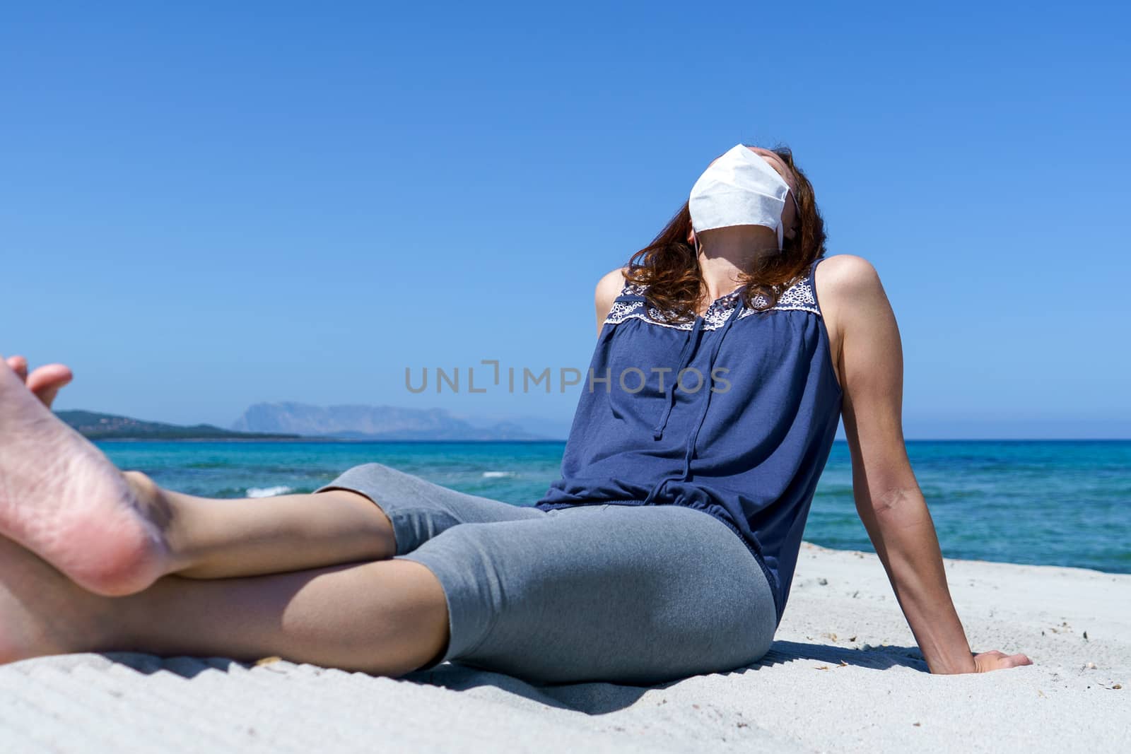 Coronavirus seaside holidays: a woman sitting on the sand at the beach look at the sun with the mask for Covid-19 pandemic