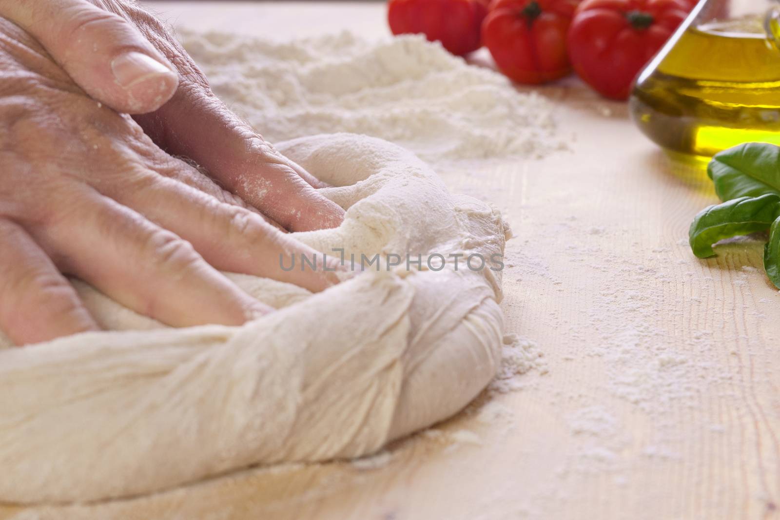 Close up of man chef hands pours and knead the pizza dough on wooden worktop and flour, extra virgin oil, tomatoes and basil in the background