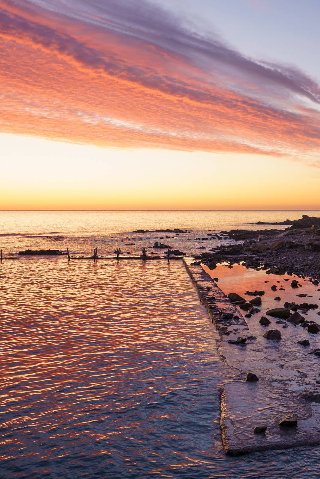 Orange and red sky with gorgeous cloud on an old abandoned sea pier in dawn or sunset with water reflections between the rocks by robbyfontanesi