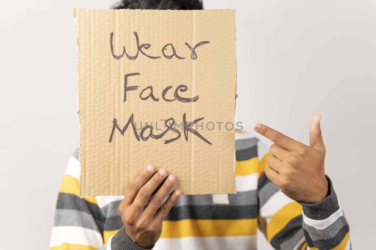 Young man holding a cardboard sign by covering his face, saying wear face mask message on isolated background to protect from coronavirus or covid-19