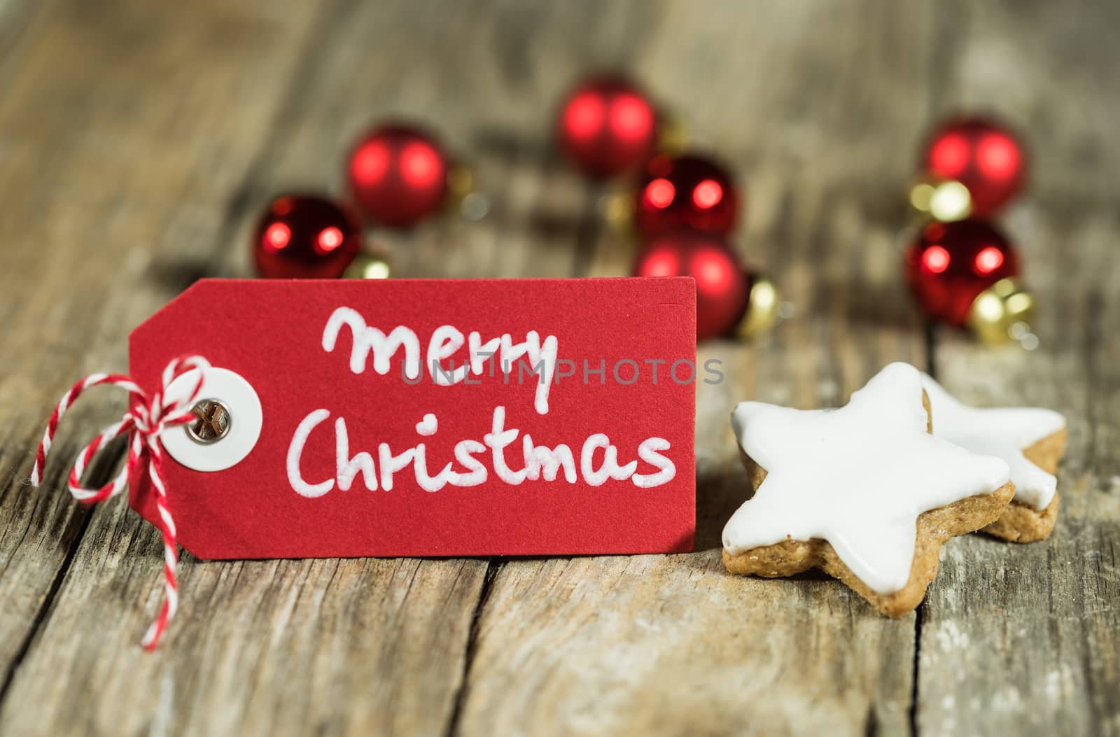 Tag with handwritten Merry Christmas greeting on wooden table with star biscuits and red baubles