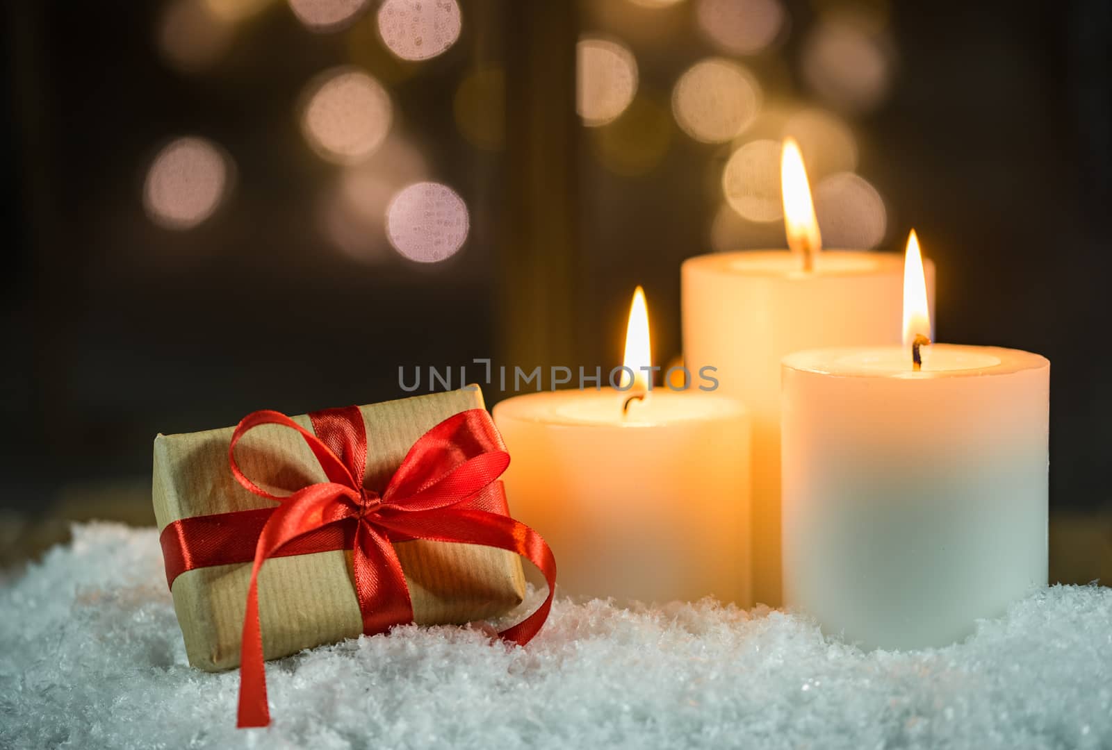 Christmas holiday decoration with three burning candles and christmas present on snow with blurred light window background