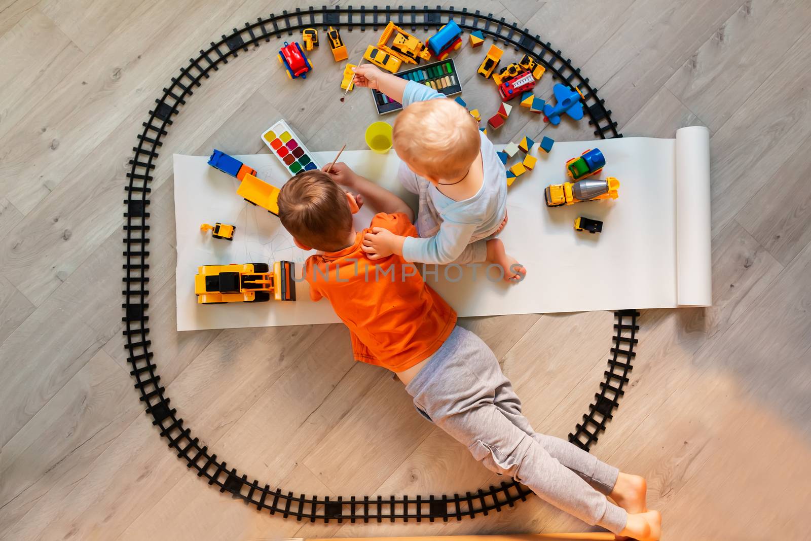 Preschool boys drawing on floor on paper, playing with educational toys - blocks, train, railroad, vehicles at home or daycare. Toys for preschool and kindergarten. Top view. Children's art and creativeness concept