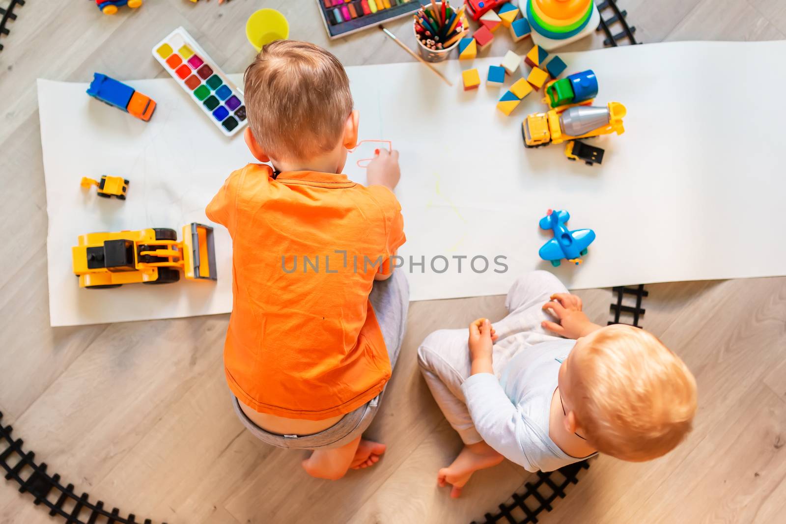 Preschool boys drawing on floor on paper, playing with educational toys - blocks, train, railroad, vehicles at home or daycare. Toys for preschool and kindergarten. Top view. Children's art and creativeness concept