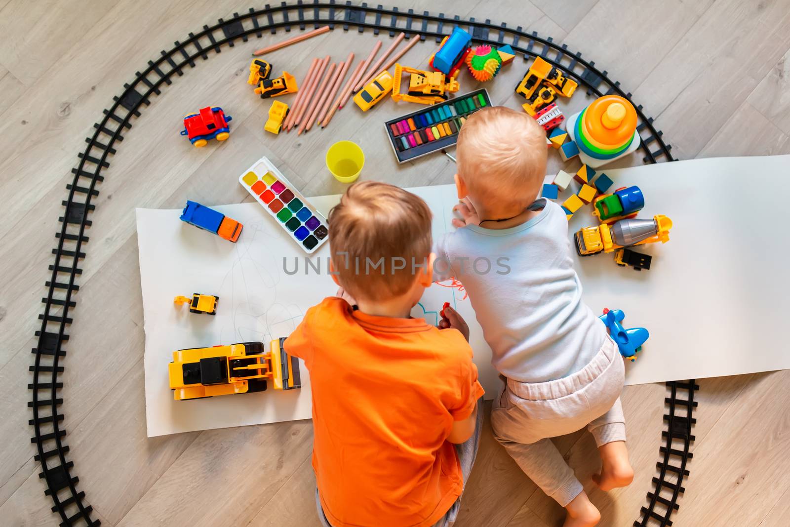 Preschool boys drawing on floor on paper, playing with educational toys - blocks, train, railroad, vehicles at home or daycare. Toys for preschool and kindergarten. Top view. Children's art and creativeness concept