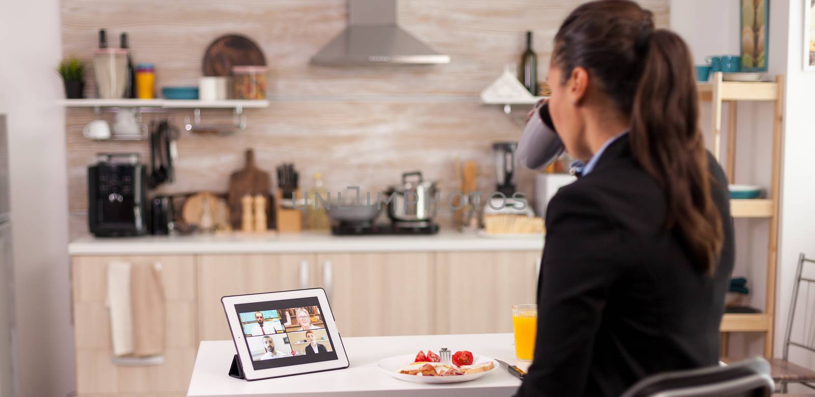 Business woman using smart device while eating breakfast in kitchen. Young freelancer at home talking on a video call with her colleagues from the office, using modern internet technology