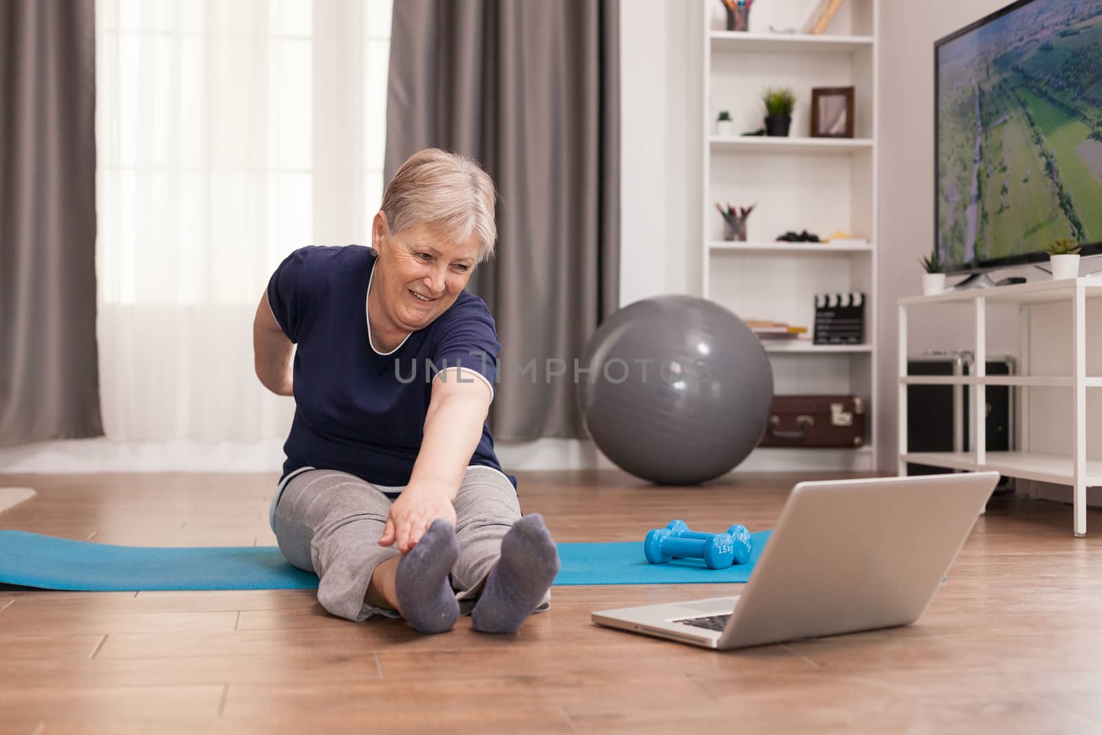 Cheerful old woman doing sports watching online lessons. Old person pensioner online internet exercise training at home sport activity with dumbbell, resistance band, swiss ball at elderly retirement age