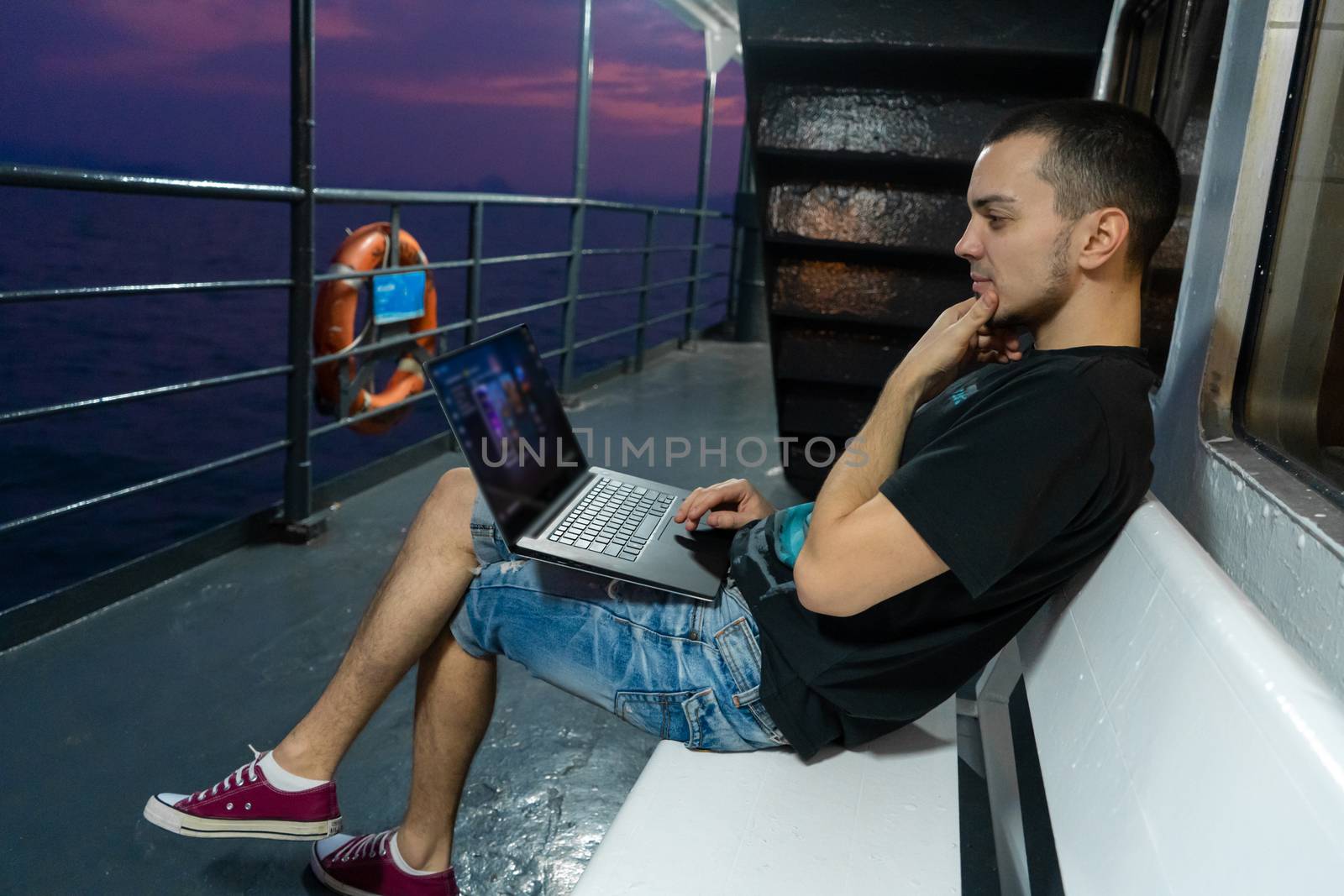 A young man works on a laptop on the deck of a ship. Remote work. Always in touch. by Try_my_best