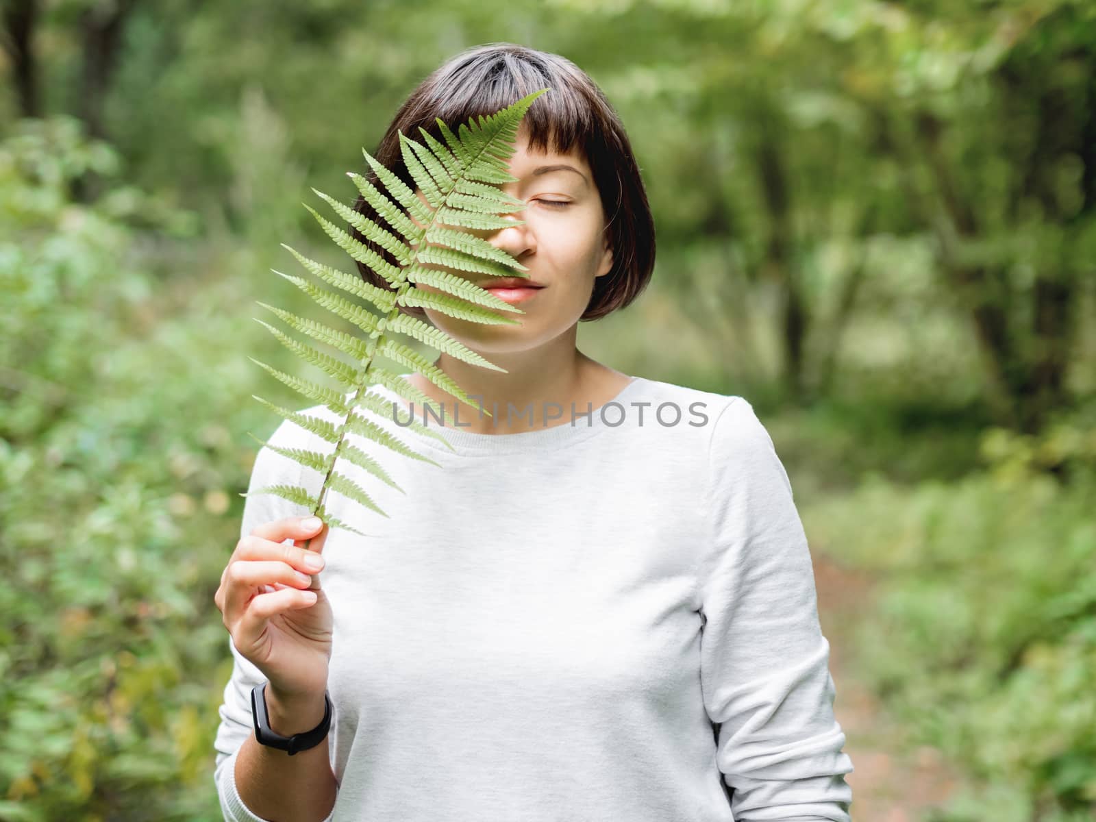 Young woman is hiding her eye with fern leaf. Symbol of life, tranquility and unity with nature. Summer in forest.