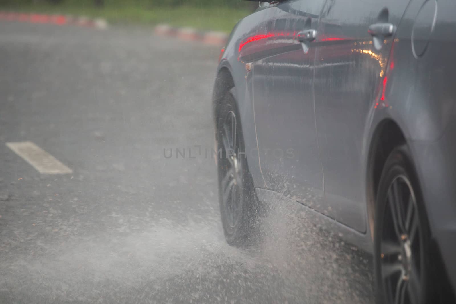 Rain water splash flow from wheels of silver car moving fast in daylight city with selective focus. Car moving on asphalt road during heavy summer storm rain.