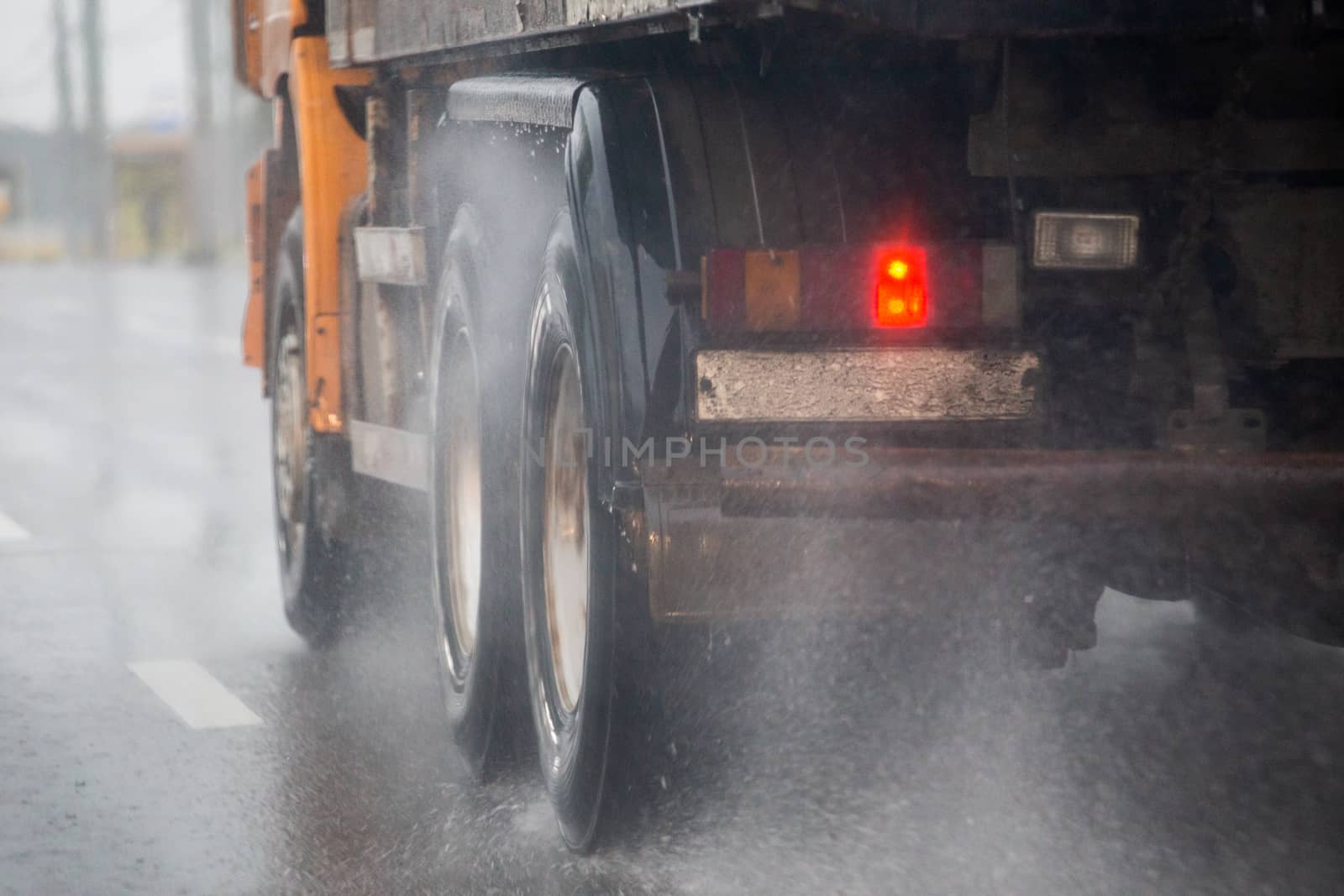 Rain water splash flow from wheels of heavy truck moving fast in daylight cit. Truck moving on asphalt road during heavy summer storm rain. Close-up with selective focus and blur.