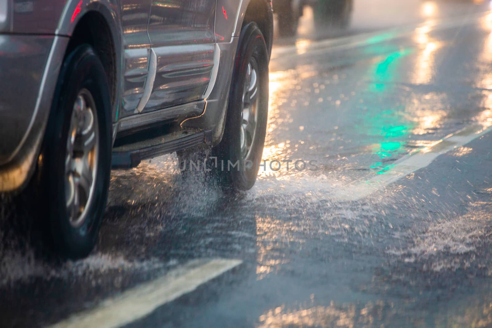 Rain water splash flow from wheels of silver car moving fast in daylight city with selective focus. Car moving on asphalt road during heavy summer storm rain.