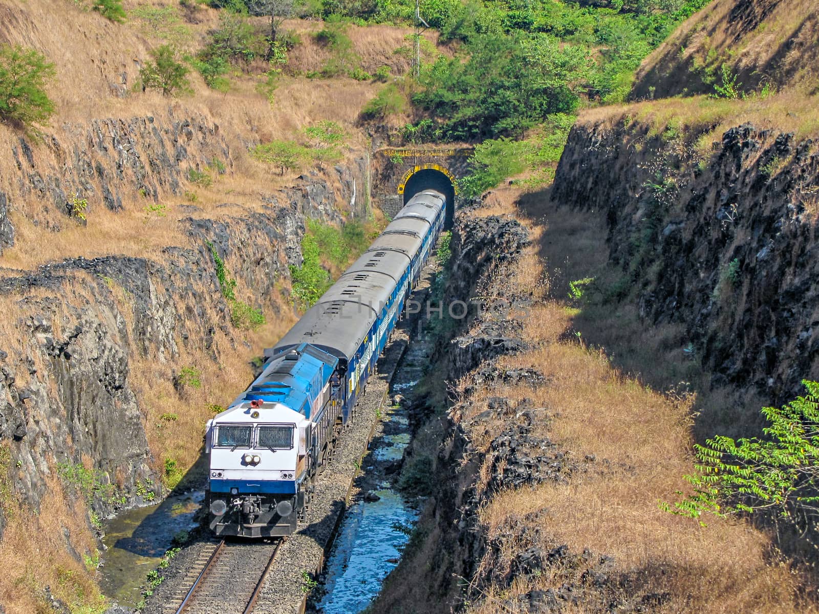 A passenger train coming out of a tunnel into deep cutting in Konkan region of Maharashtra, India. by lalam