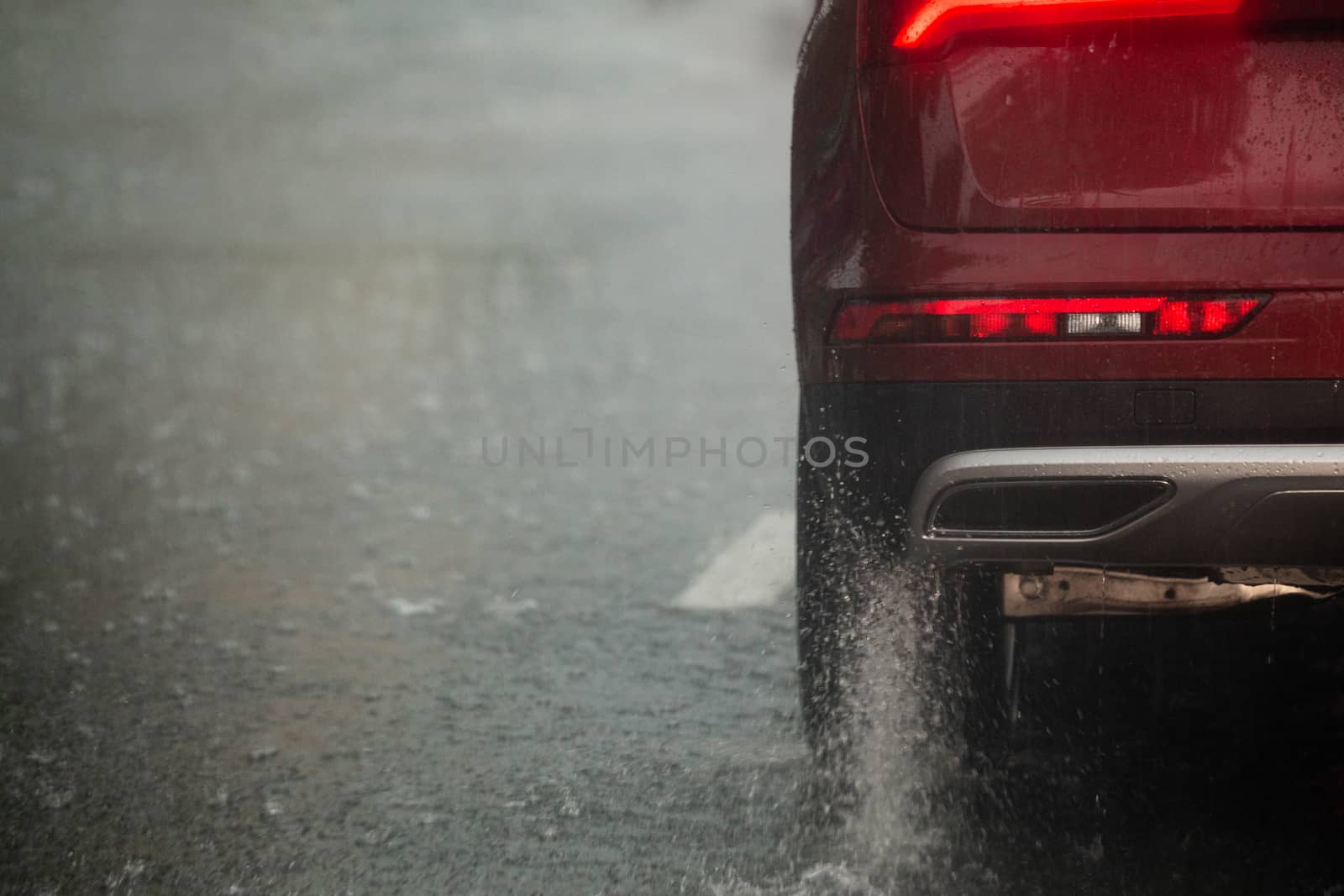 Rain water splash flow from wheels of red car moving fast in daylight city with selective focus. Car moving on asphalt road during heavy summer storm rain.