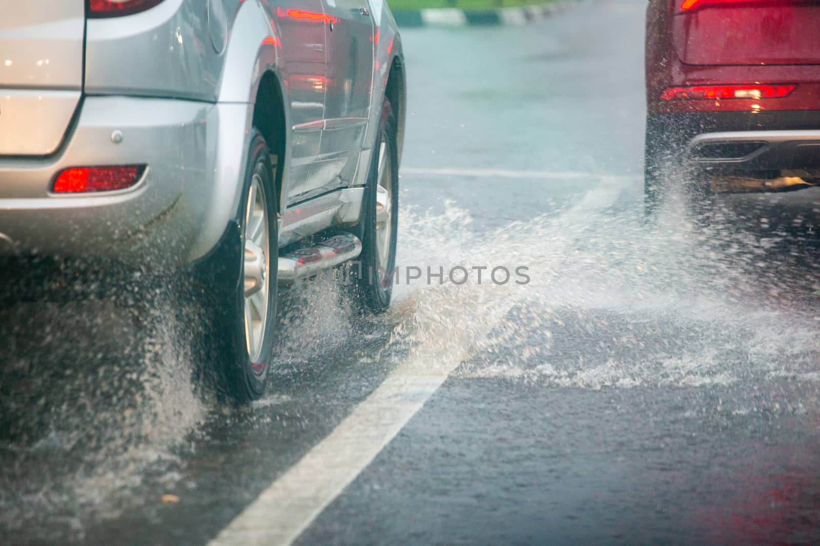 Rain water splash flow from wheels of silver and red cars moving fast in daylight city with selective focus. Car moving on asphalt road during heavy summer storm rain.