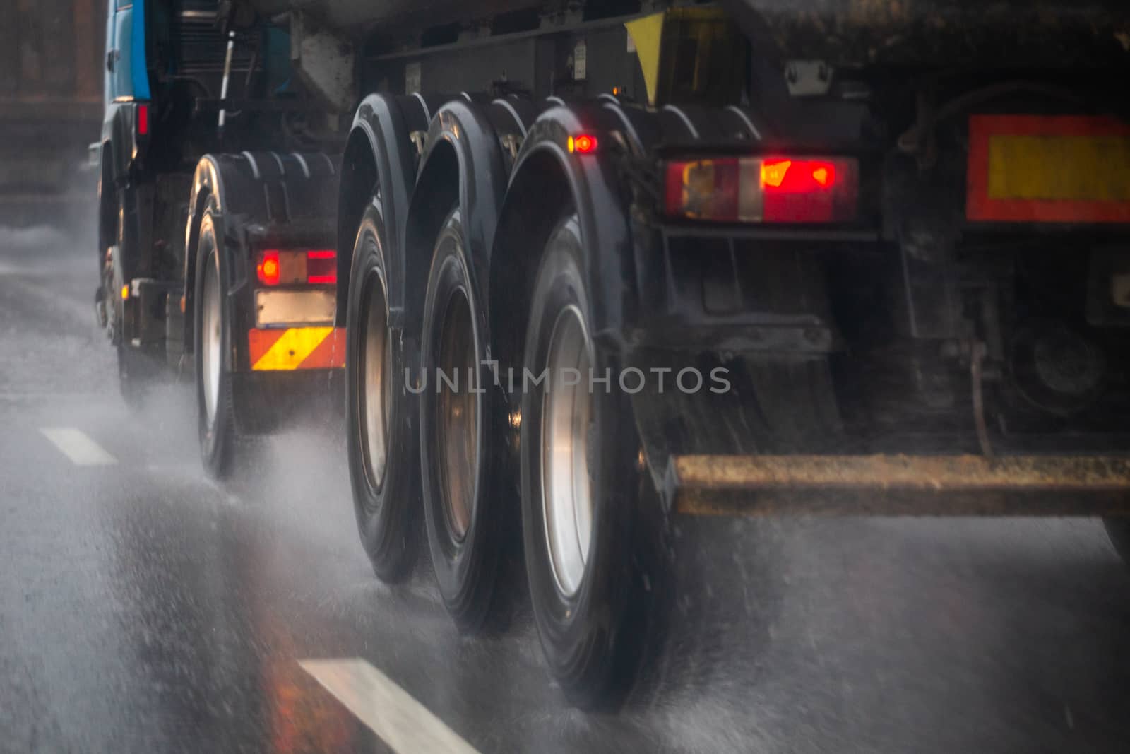 Rain water splash flow from wheels of heavy truck moving fast in daylight cit. Truck moving on asphalt road during heavy summer storm rain. Close-up with selective focus and blur.