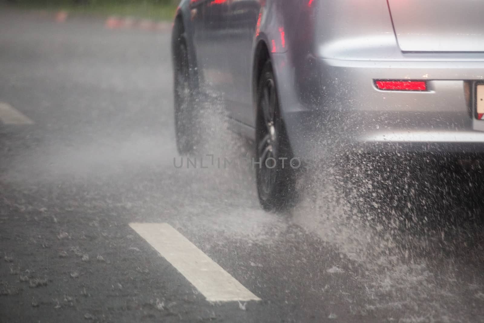 Rain water splash flow from wheels of silver car moving fast in daylight city with selective focus. Car moving on asphalt road during heavy summer storm rain.