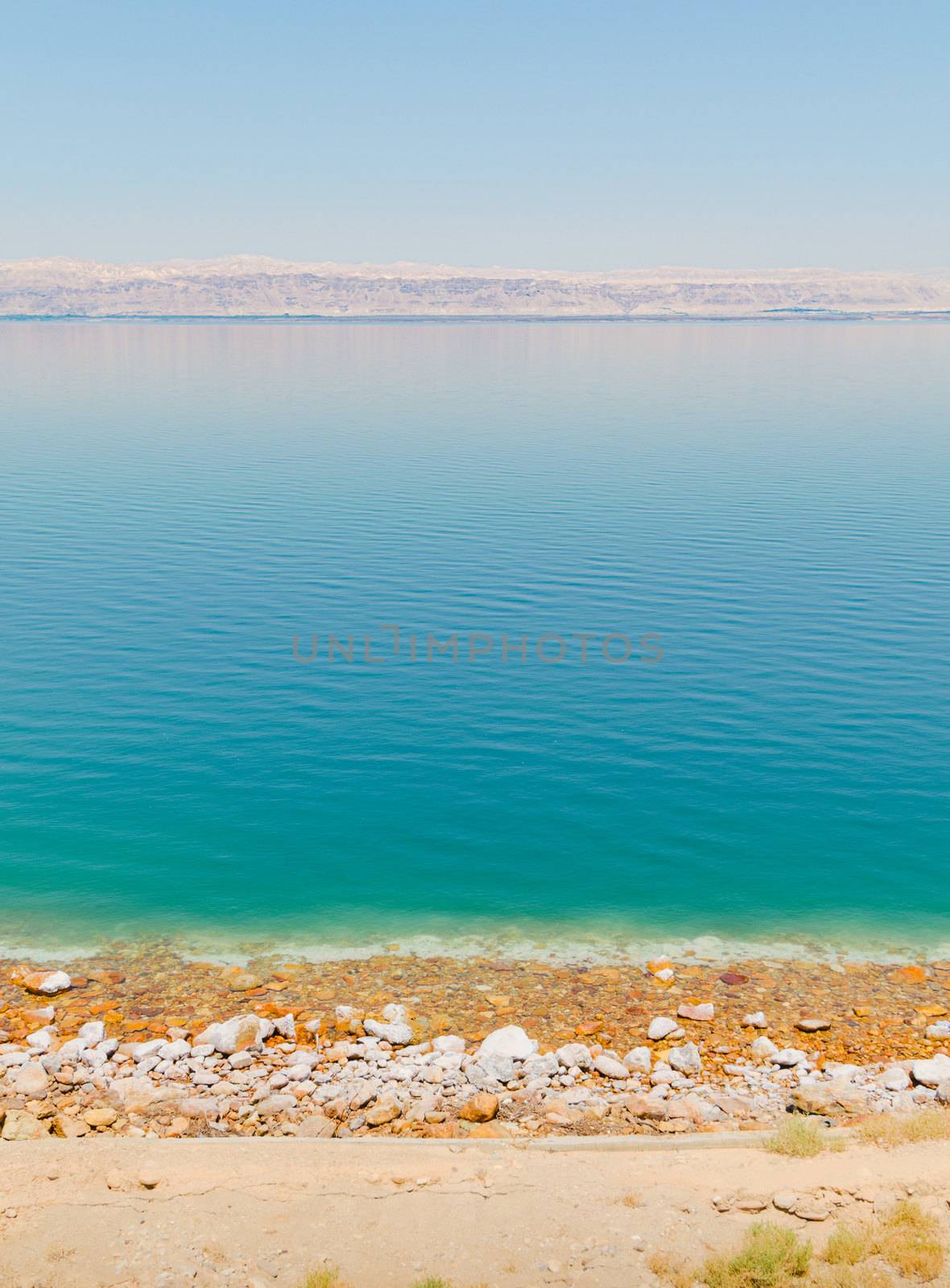 Vertical landscape view of the Dead sea shore, with salt crust and pebble beach near a tourist resort, and Israel territories mountain line on the horizon, from Madaba, Jordan, Middle East