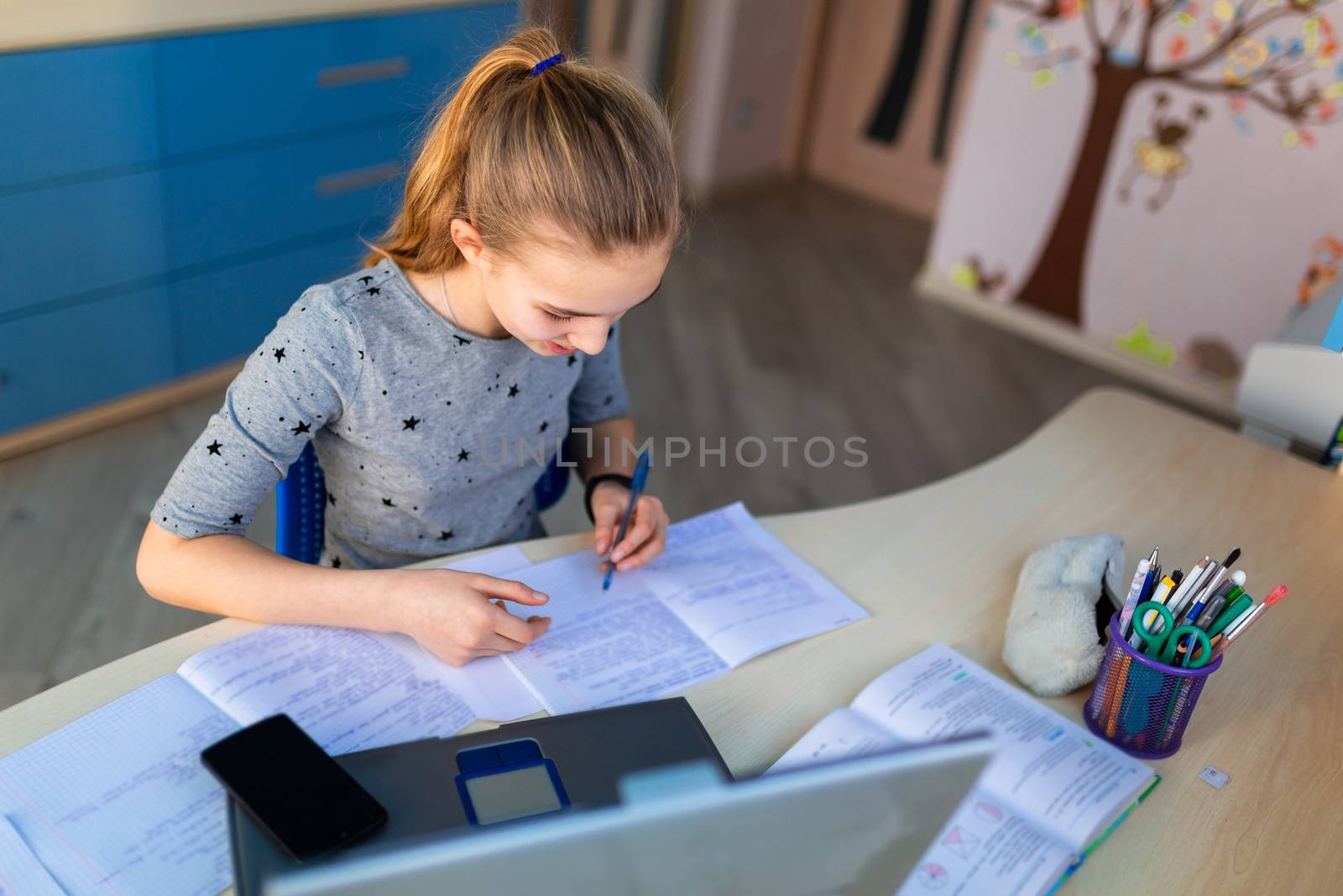 Beautiful young school girl working at home in her room with a laptop and class notes studying in a virtual class. Distance education and learning concept during quarantine by Len44ik