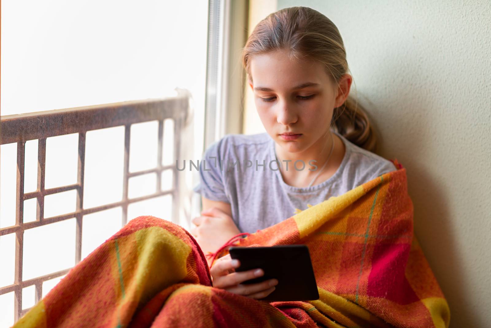 Sad little girl sitting on the window with e-book or tablet reading and studying at home isolated. Coronavirus quarantine distance education concept.
