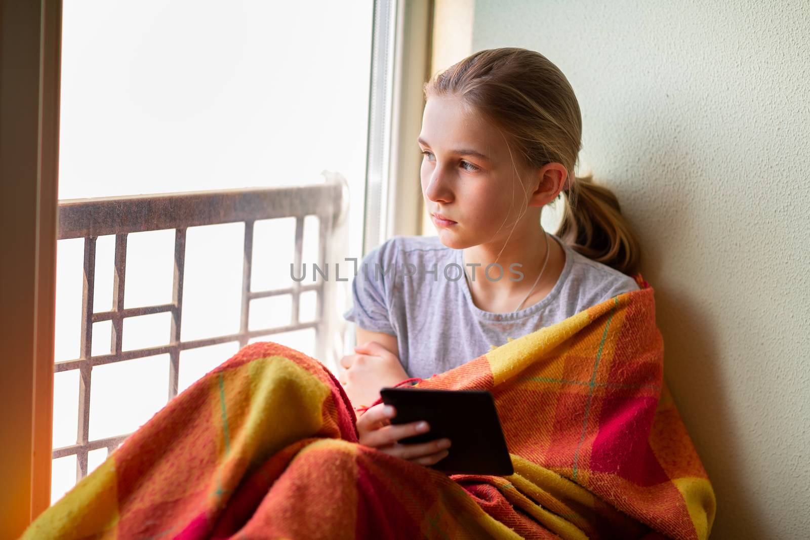 Sad little girl sitting on the window with e-book or tablet reading and studying at home isolated. Coronavirus quarantine distance education concept.